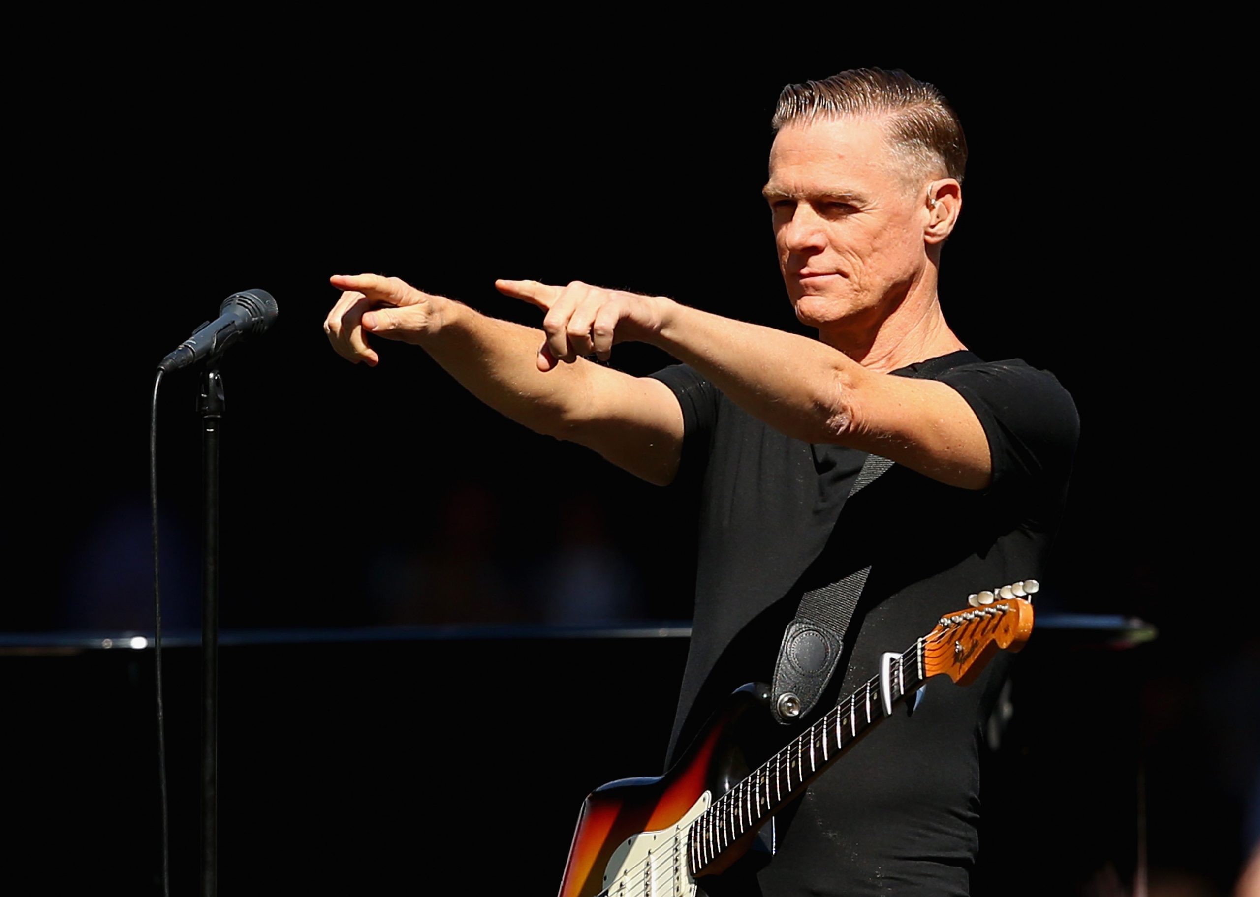 Bryan adams pointing fingers at the audience behind a microphone wearing a black tee with his guita...