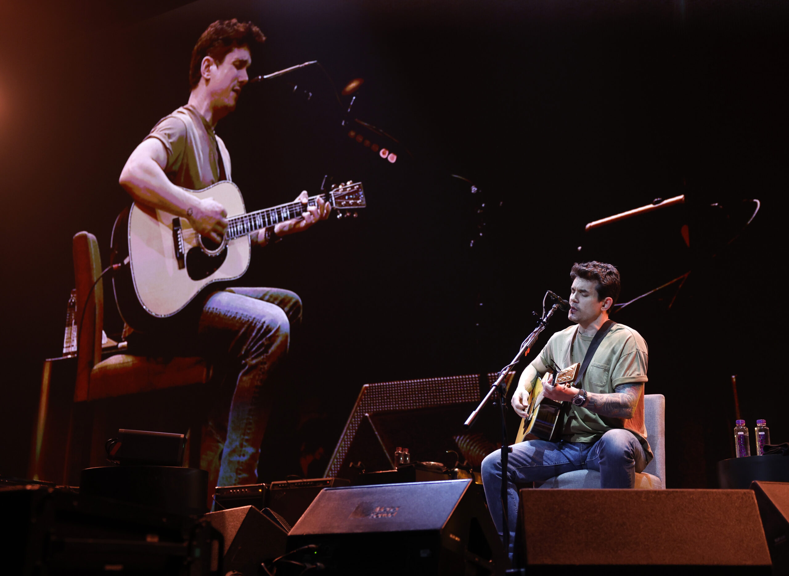 John Mayer performing on stage while seated with a projection of himself on the screen behind him...